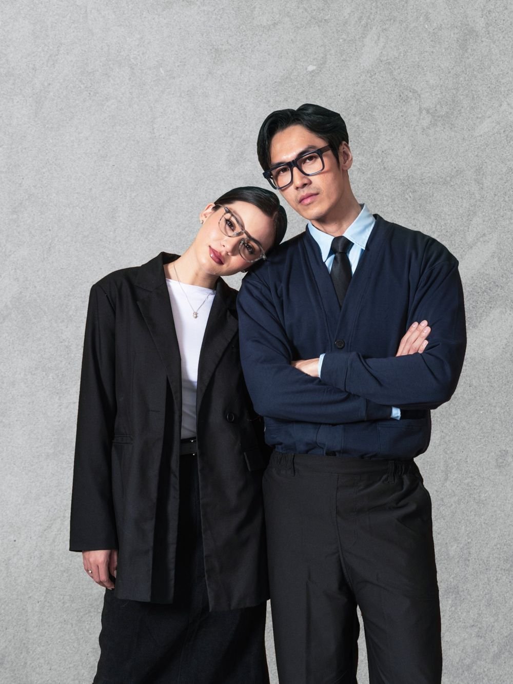 Couple in glasses posing against a concrete wall